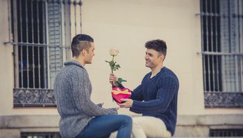 young cool gay men couple on street with rose flower and heart shape box present giving gift celebrating valentines day in love on street urban background